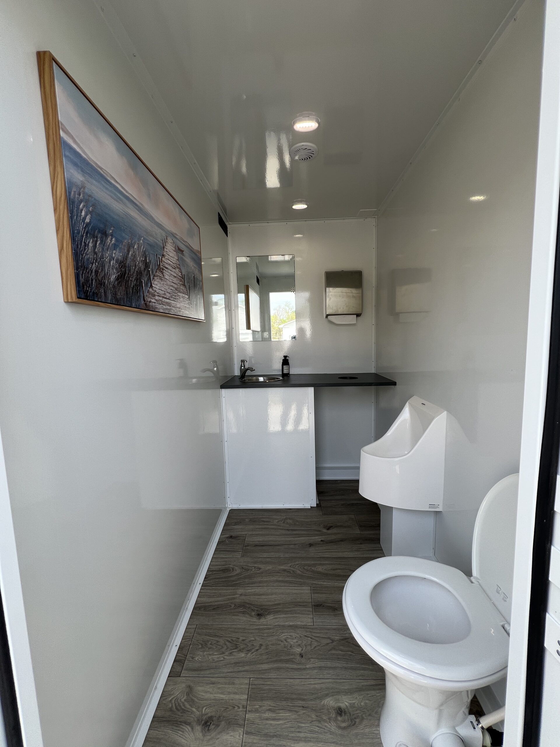 Luxury restroom trailer men's stall featuring toilet, urinal, sink, framed coastal artwork, and wood-look flooring – High Cotton Comfort Co