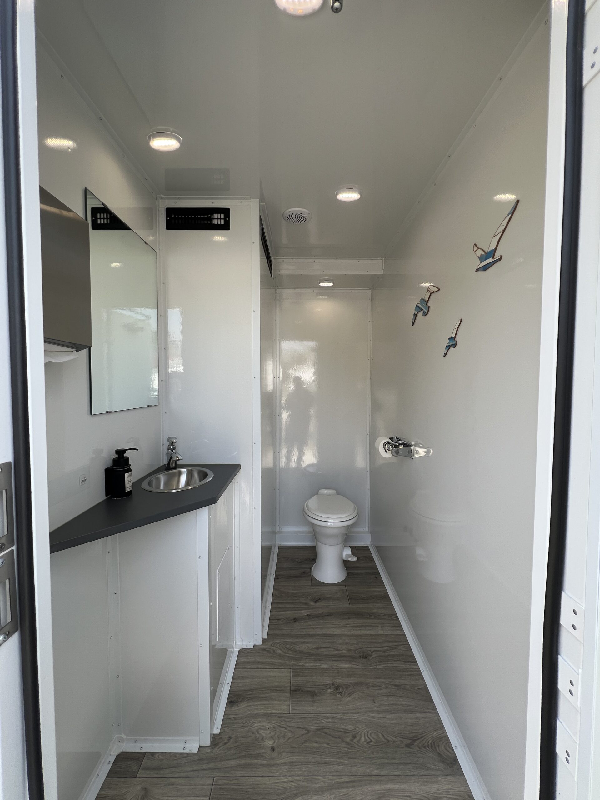 Luxury restroom trailer women's stall interior with toilet, sink, mirror, wood-style flooring, and bright LED lighting – High Cotton Comfort Co Charleston