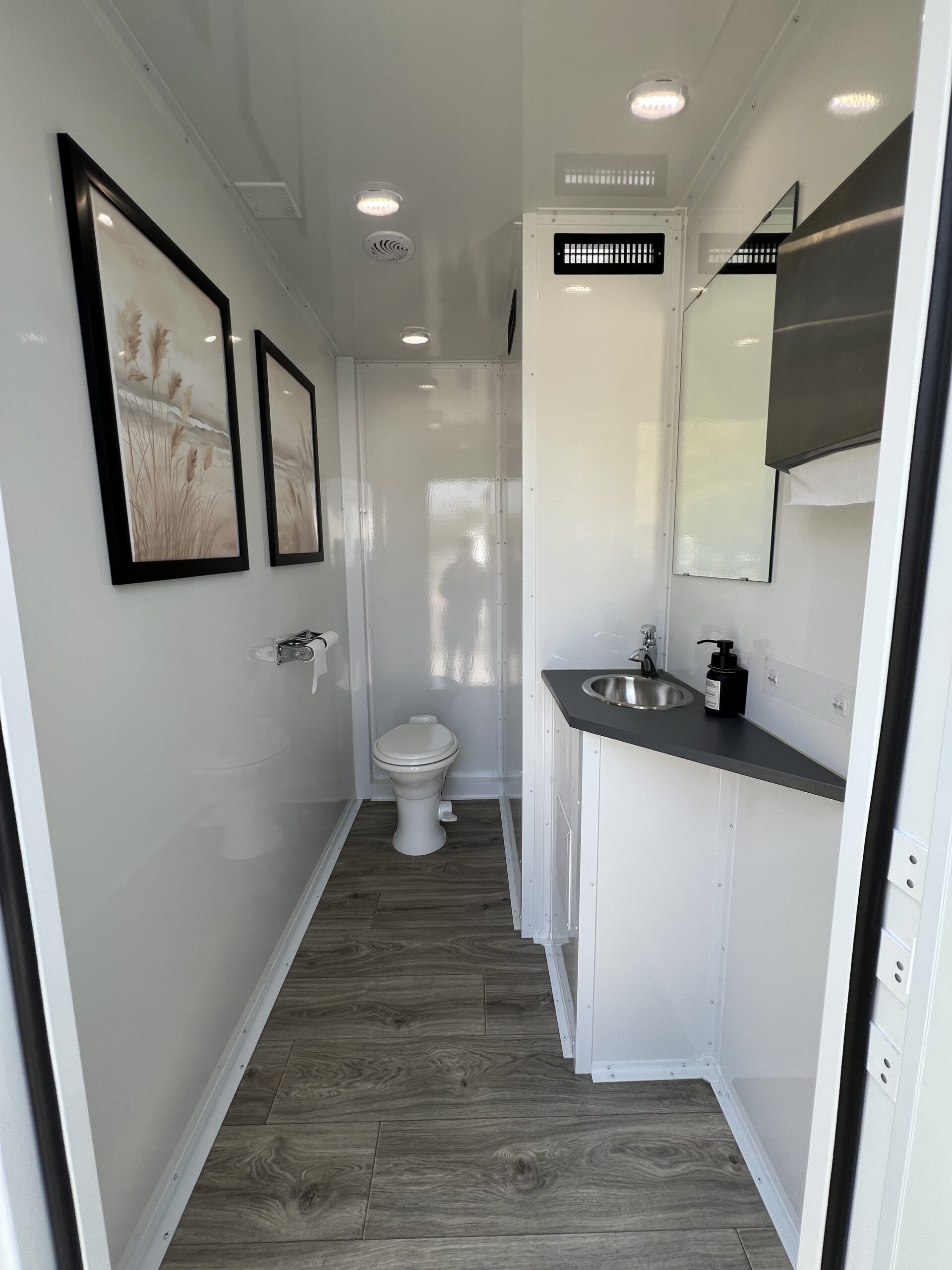 Upscale restroom trailer stall with toilet, sink vanity, mirror, framed wall art, and wood-style flooring – High Cotton Comfort Co 3-stall trailer
